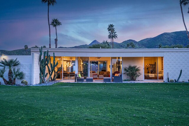 back house at dusk with an outdoor hangout area, ceiling fan, a mountain view, and a lawn