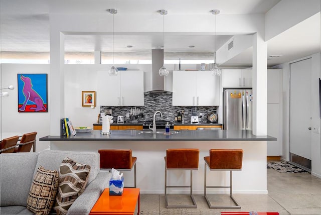 kitchen with white cabinetry, hanging light fixtures, wall chimney range hood, a breakfast bar, and stainless steel refrigerator