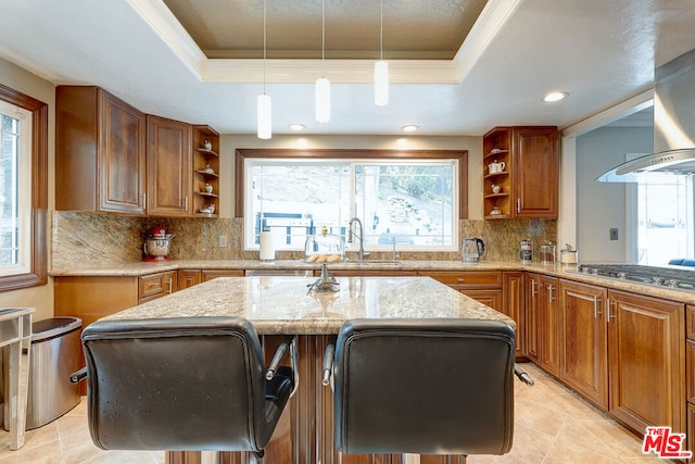 kitchen with stainless steel gas stovetop, a kitchen island, decorative backsplash, a tray ceiling, and light stone counters