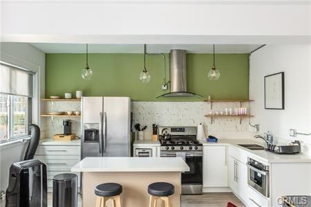 kitchen featuring hanging light fixtures, appliances with stainless steel finishes, wall chimney range hood, and white cabinetry