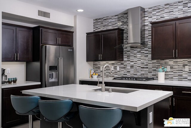 kitchen featuring appliances with stainless steel finishes, wall chimney exhaust hood, a kitchen bar, an island with sink, and tasteful backsplash