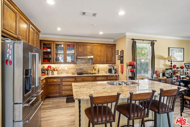 kitchen featuring stainless steel appliances, sink, light stone counters, a center island with sink, and crown molding