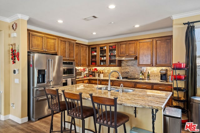 kitchen with an island with sink, stainless steel appliances, light stone countertops, ornamental molding, and light hardwood / wood-style flooring