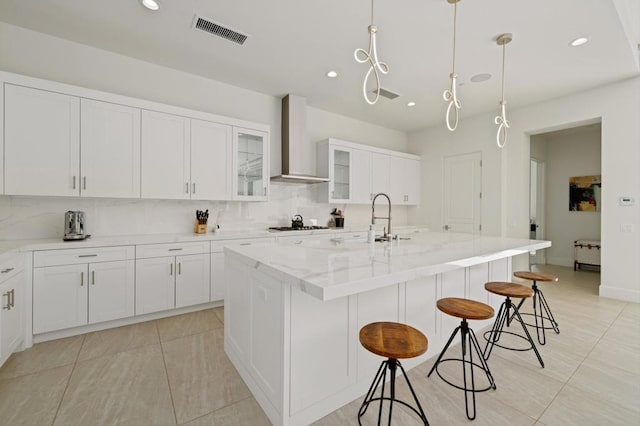 kitchen featuring a center island with sink, a breakfast bar area, pendant lighting, light stone countertops, and wall chimney exhaust hood