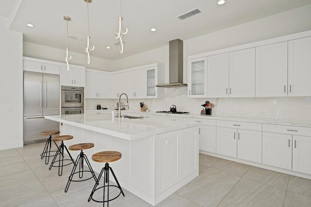 kitchen featuring appliances with stainless steel finishes, decorative light fixtures, wall chimney range hood, sink, and a kitchen island with sink