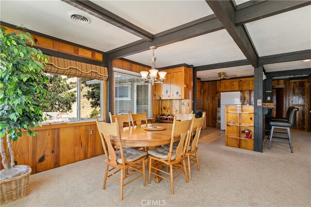 dining room featuring beam ceiling, light colored carpet, wood walls, and ceiling fan with notable chandelier