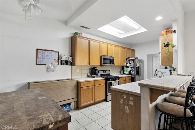 kitchen featuring tile counters, stainless steel appliances, decorative backsplash, a kitchen breakfast bar, and light tile patterned floors