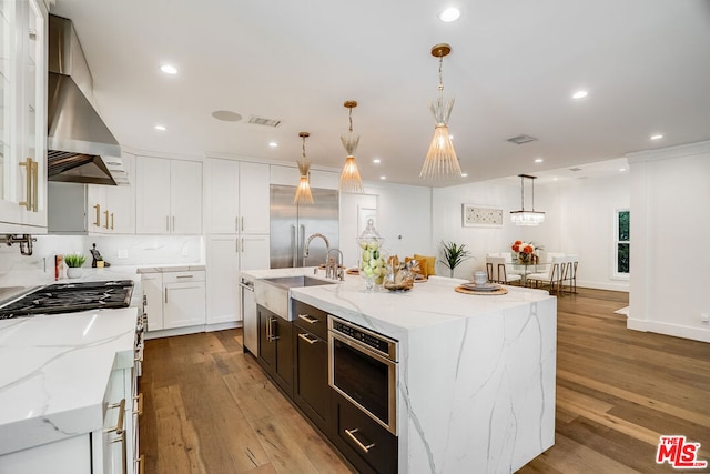 kitchen featuring wall chimney exhaust hood, light stone counters, hanging light fixtures, a large island with sink, and white cabinets