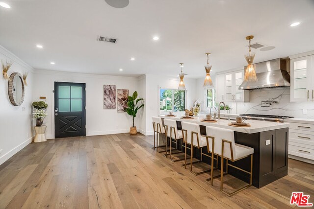 kitchen featuring a kitchen bar, white cabinetry, decorative light fixtures, a center island with sink, and wall chimney range hood