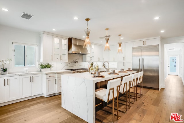 kitchen featuring appliances with stainless steel finishes, a kitchen island with sink, white cabinets, decorative light fixtures, and wall chimney exhaust hood