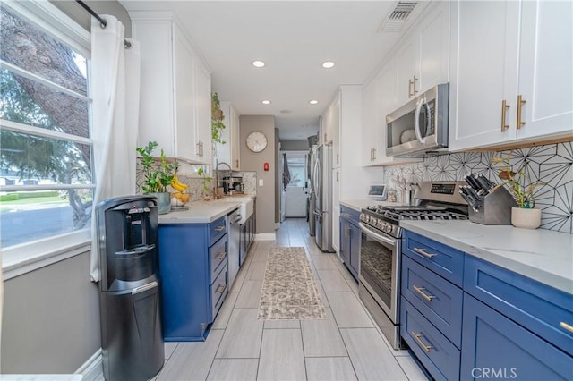 kitchen featuring decorative backsplash, appliances with stainless steel finishes, blue cabinetry, white cabinets, and light stone counters