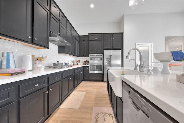 kitchen featuring light wood finished floors, dark cabinets, stainless steel appliances, under cabinet range hood, and a sink