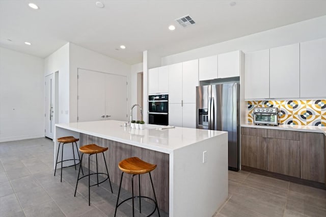 kitchen with white cabinets, black double oven, stainless steel fridge with ice dispenser, a kitchen island with sink, and a breakfast bar area