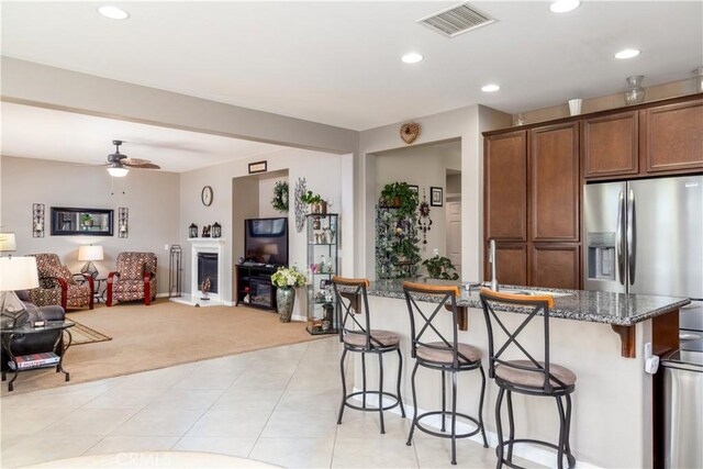 kitchen with ceiling fan, light colored carpet, stainless steel refrigerator with ice dispenser, an island with sink, and dark stone counters