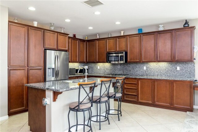 kitchen featuring dark stone countertops, a kitchen island with sink, a kitchen breakfast bar, stainless steel appliances, and light tile patterned floors