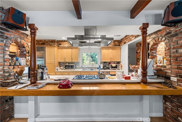 kitchen featuring beam ceiling, island exhaust hood, light brown cabinetry, and brick wall