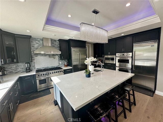 kitchen featuring wall chimney exhaust hood, decorative light fixtures, built in appliances, a tray ceiling, and a center island with sink