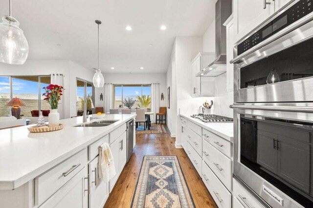 kitchen featuring white cabinetry, wall chimney range hood, sink, hanging light fixtures, and a center island with sink