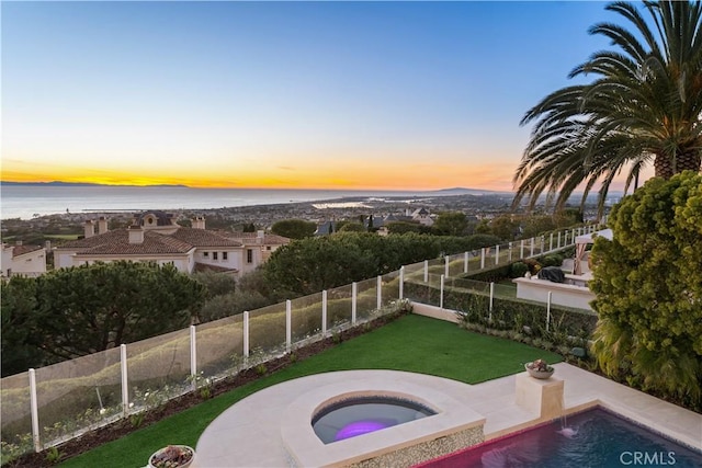 pool at dusk with a water view, an in ground hot tub, and a yard