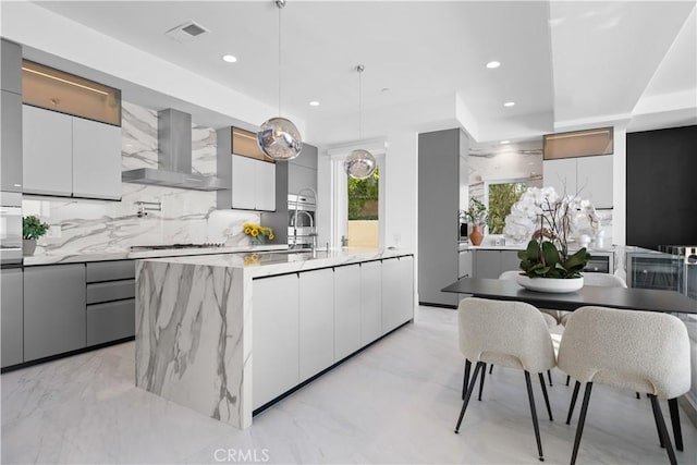 kitchen featuring pendant lighting, wall chimney exhaust hood, gray cabinetry, tasteful backsplash, and a center island with sink