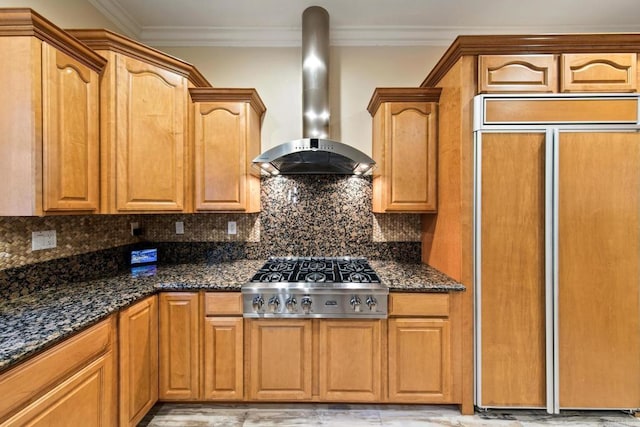 kitchen with paneled built in refrigerator, wall chimney range hood, ornamental molding, dark stone countertops, and stainless steel gas stovetop