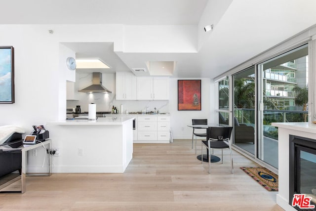 kitchen featuring wall chimney range hood, white cabinets, kitchen peninsula, and light wood-type flooring