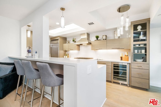 kitchen with light brown cabinetry, wall chimney exhaust hood, beverage cooler, and decorative light fixtures