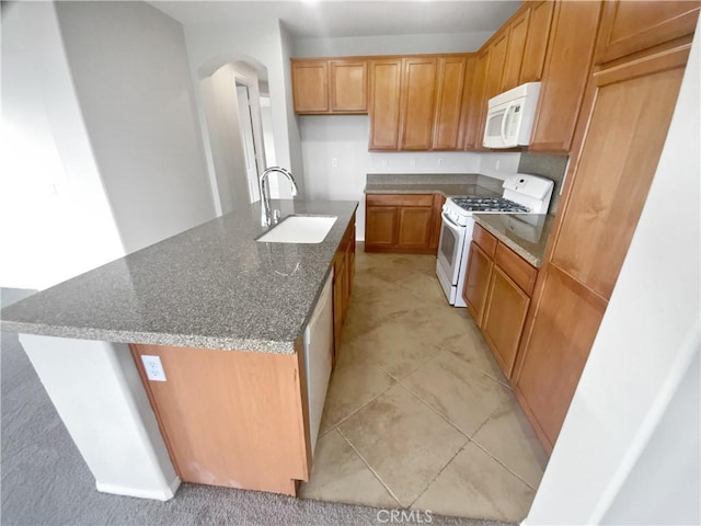 kitchen with arched walkways, a kitchen island with sink, white appliances, a sink, and dark stone countertops