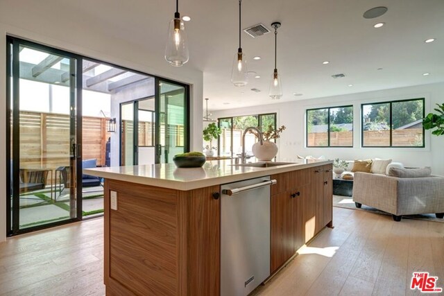 kitchen with light hardwood / wood-style floors, stainless steel dishwasher, a center island with sink, sink, and hanging light fixtures