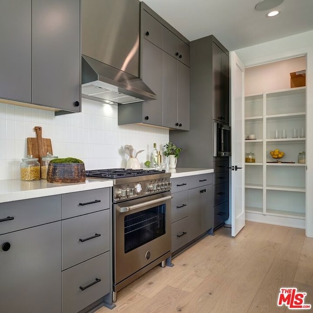 kitchen with high end stainless steel range, wall chimney exhaust hood, gray cabinetry, backsplash, and light hardwood / wood-style flooring