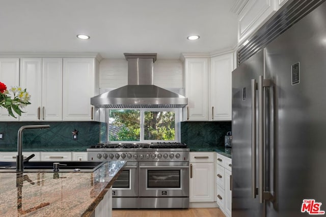 kitchen with premium appliances, white cabinets, wall chimney exhaust hood, dark stone counters, and backsplash