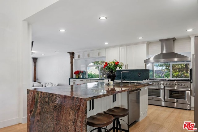 kitchen with appliances with stainless steel finishes, a center island with sink, dark stone counters, and wall chimney range hood