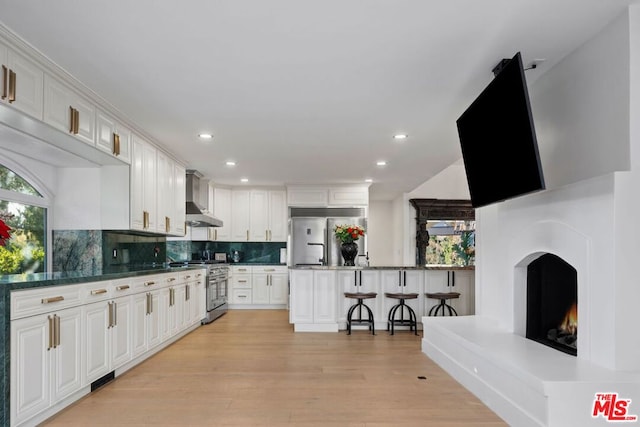 kitchen featuring a kitchen breakfast bar, stainless steel appliances, wall chimney exhaust hood, and white cabinets