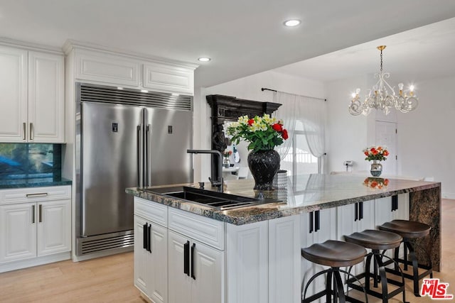 kitchen with built in refrigerator, white cabinets, an inviting chandelier, an island with sink, and sink