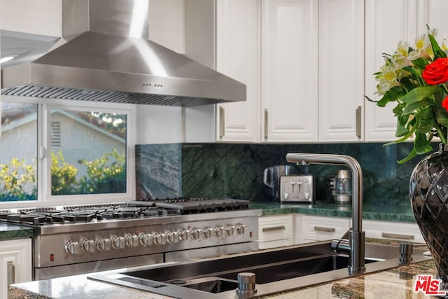 kitchen with tasteful backsplash, wall chimney range hood, high end stainless steel range oven, white cabinets, and dark stone counters