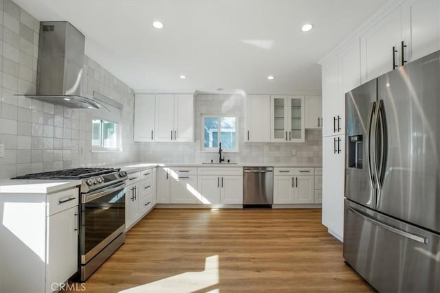 kitchen with white cabinets, appliances with stainless steel finishes, wall chimney range hood, backsplash, and light wood-type flooring