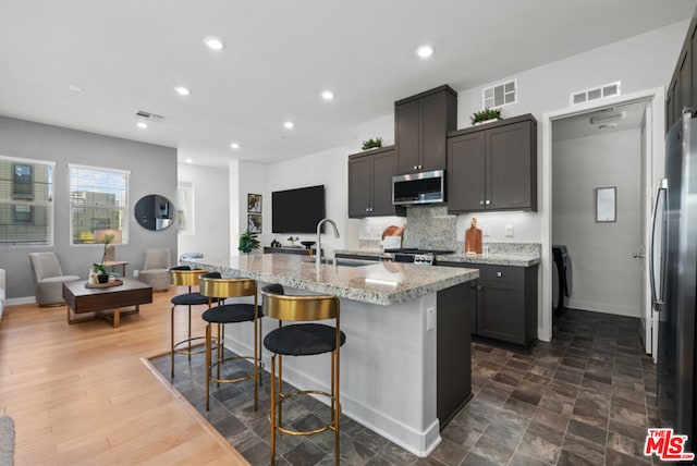 kitchen with backsplash, a center island with sink, sink, appliances with stainless steel finishes, and light stone counters