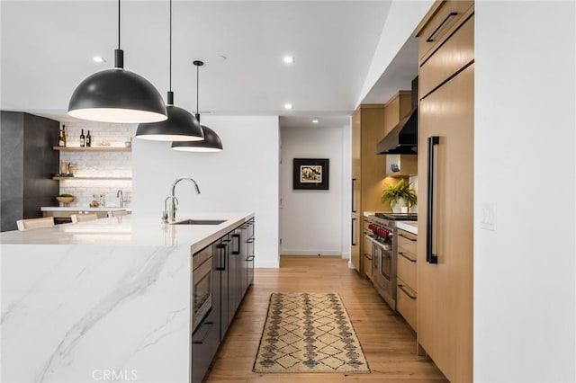 kitchen featuring appliances with stainless steel finishes, wall chimney range hood, light stone countertops, pendant lighting, and sink