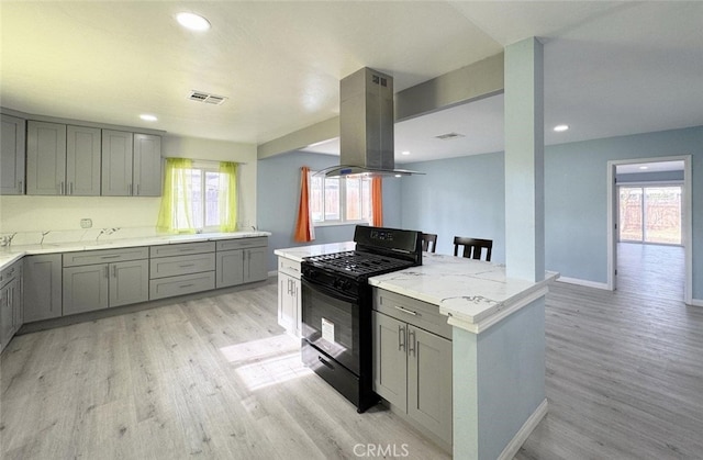 kitchen with black gas range oven, island exhaust hood, light stone counters, gray cabinetry, and light hardwood / wood-style floors