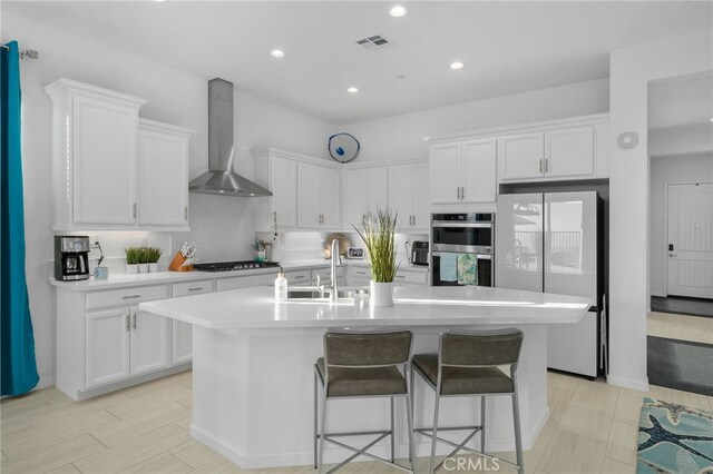 kitchen with white cabinetry, wall chimney range hood, stainless steel appliances, an island with sink, and sink