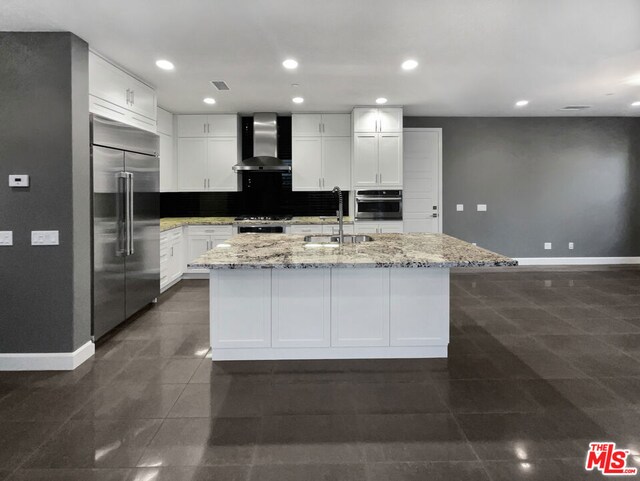 kitchen featuring a kitchen island with sink, wall chimney range hood, stainless steel appliances, and white cabinetry