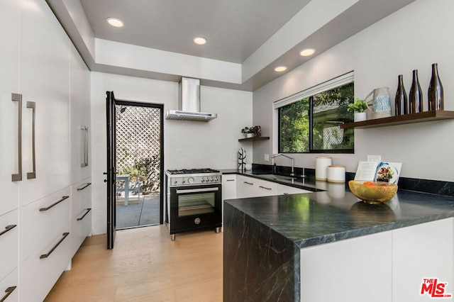 kitchen with wall chimney range hood, sink, light hardwood / wood-style flooring, stainless steel range, and white cabinets