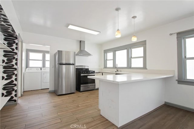 kitchen featuring appliances with stainless steel finishes, wall chimney range hood, washing machine and dryer, a peninsula, and light wood-style floors