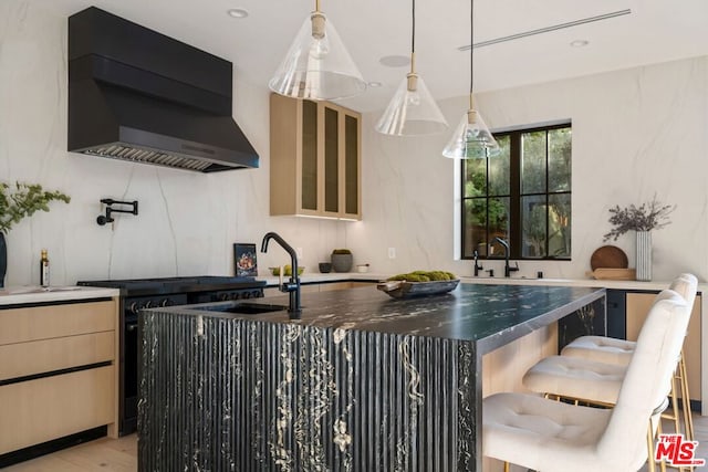 kitchen featuring black stove, a center island with sink, sink, light brown cabinets, and wall chimney exhaust hood