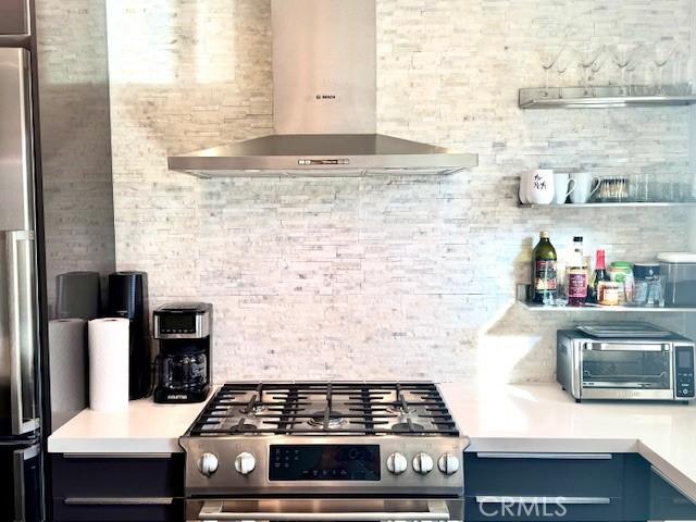 kitchen featuring blue cabinetry, tasteful backsplash, stove, stainless steel refrigerator, and wall chimney range hood