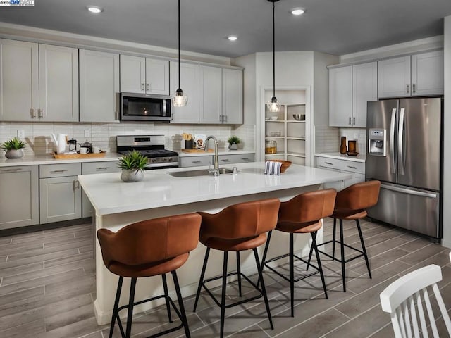 kitchen featuring a center island with sink, gray cabinets, appliances with stainless steel finishes, hanging light fixtures, and sink