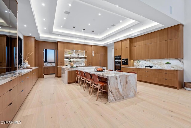 kitchen with a kitchen bar, a tray ceiling, hanging light fixtures, light stone countertops, and a large island