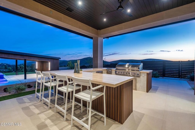 patio terrace at dusk with a mountain view, ceiling fan, exterior kitchen, a grill, and an outdoor bar