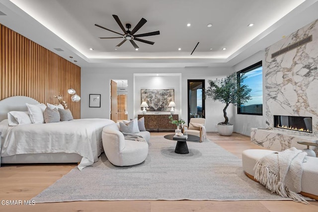 bedroom with ceiling fan, a premium fireplace, a tray ceiling, and light hardwood / wood-style floors