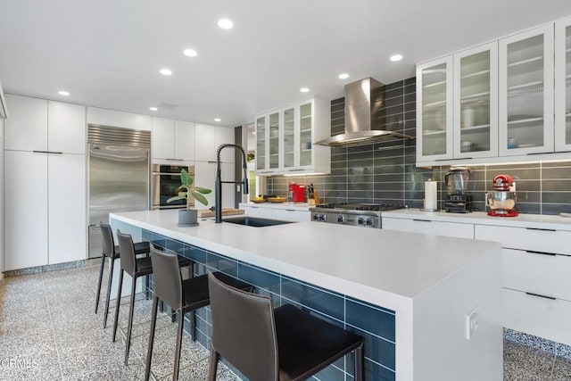 kitchen featuring white cabinets, wall chimney exhaust hood, stainless steel appliances, a kitchen breakfast bar, and a kitchen island with sink